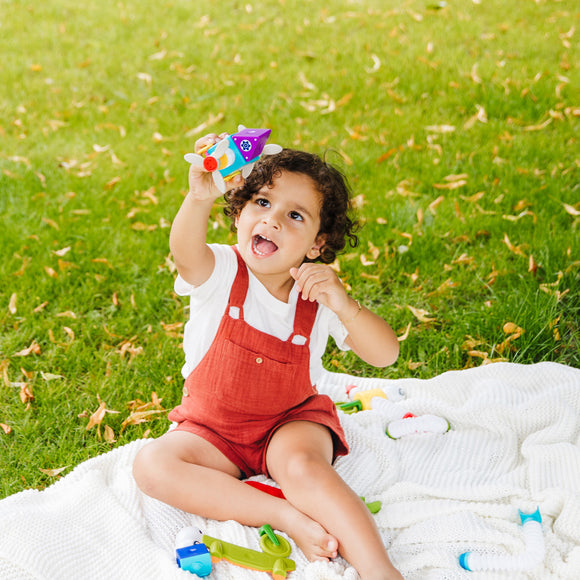 A joyful child with curly hair, wearing a white shirt and orange overalls, sitting on a white blanket in a grassy area. The child is holding a colorful toy airplane aloft, showcasing their excitement. Surrounding the child are various other colorful toys, encouraging imaginative play and creativity.