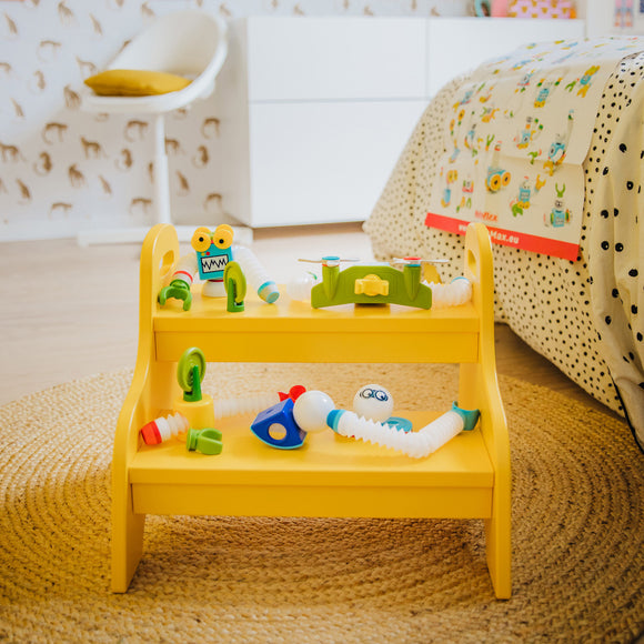 The Smartmax Roboflex Large set displayed on a stylish yellow shelf. It includes various colorful flexible components like tubes, connectors, and playful characters. The background features a patterned wall and a neat bed, creating a vibrant playroom atmosphere.