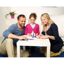 Three people, a man, a woman, and a girl, are sitting around a white table playing with the Tangoes Classic mechanical puzzle by SmartGames. The girl, wearing a pink sweater, is focused on a colorful arrangement of pieces while the adults assist her with smiles. The setting is bright and inviting, featuring a plant in the background and a cozy rug on the floor, emphasizing a fun family activity.