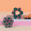 A colorful set of Speks Stripes Nebula magnets arranged on a surface. It features a green and purple structure in a flower shape and a round shape, showcasing the versatility and creativity allowed by these magnetic pieces. Nearby, there are colored pencils and a notebook, enhancing the artistic theme.