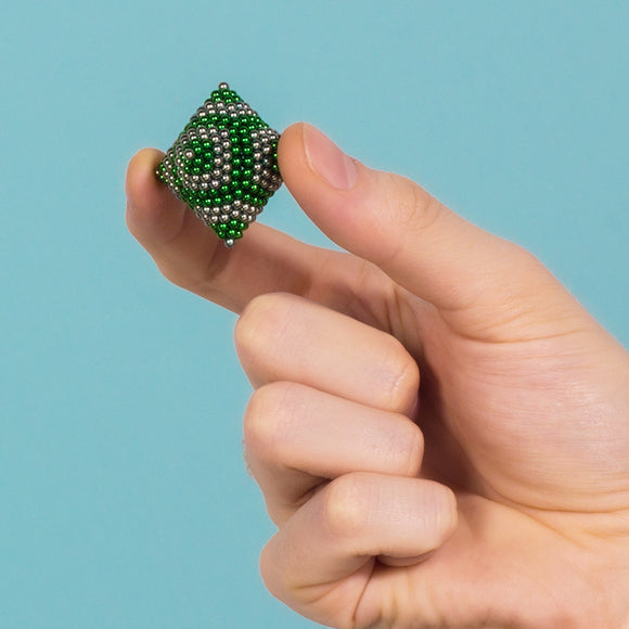 A hand holding a small, spherical structure made of metallic beads in green and silver colors, showcasing the Speks Stripes Third Rock fidget toy against a light blue background.