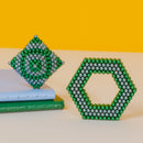 A vibrant display of Speks Stripes Third Rock featuring two geometric shapes made of colorful magnetic balls. One shape is a spiraled diamond, and the other is a hexagon, both showcasing shades of green and silver. They are placed on top of a light blue and green notebook against a bright yellow background, highlighting their playful design and tactile nature.