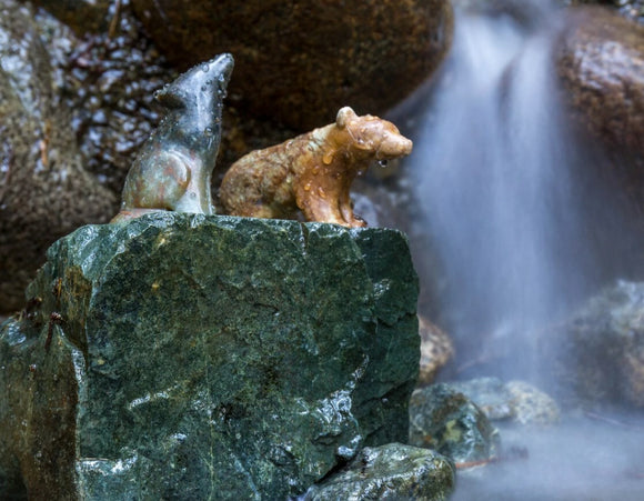 A stone carving featuring a bear and a wolf, displayed atop a green rock in a serene waterfall setting. The bear is in a seated position, while the wolf is standing with its head held high. Water droplets glisten on the smooth surface of the carvings, accentuating their details. The natural scenery adds a tranquil ambiance, perfect for nature lovers and collectors.