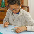 A child focused on carving a stone figure of a bear and wolf using a small hand tool. The child is wearing a light gray cable-knit sweater and glasses, sitting at a table with a blue cloth. In the background, there is a wooden dresser, adding warmth to the setting.