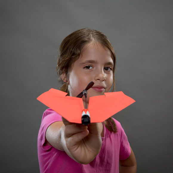 A young girl wearing a pink shirt holds the PowerUp 2.0 electric plane in front of her. The plane is brightly colored in orange and features a small propeller. The background is a neutral gray, emphasizing the child's proud expression as she presents her DIY model.