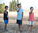 Three children outdoors in a field, smiling and holding homemade toy airplanes made from the PowerUp 2.0 Elec Plane DIY kit by TAILOR TOYS. The kids are dressed in casual summer clothes, showcasing their colorful planes against a scenic backdrop of trees and a clear sky.