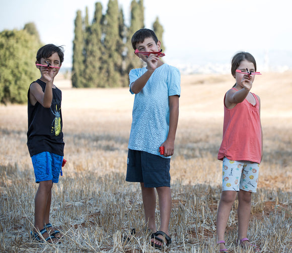Three children outdoors in a field, smiling and holding homemade toy airplanes made from the PowerUp 2.0 Elec Plane DIY kit by TAILOR TOYS. The kids are dressed in casual summer clothes, showcasing their colorful planes against a scenic backdrop of trees and a clear sky.