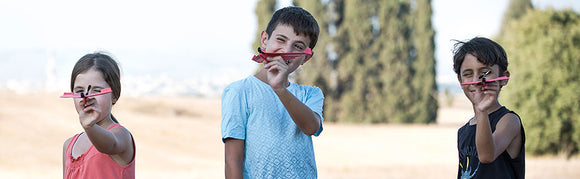 Three children outdoors, joyfully holding up their DIY PowerUp 2.0 Electric Plane kits. Each child is showcasing their colorful planes, demonstrating creativity and excitement. The background features a lush, green landscape, emphasizing outdoor play.