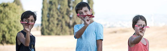 Three children outdoors holding their handmade PowerUp 2.0 electric planes, smiling and excited about their DIY creations. The background features greenery and an open field, showcasing their joy in play and creativity.