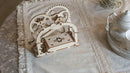 A wooden 3D mechanical puzzle kit by UGears. The intricate design features gears and a box mechanism, showcasing craftsmanship and engineering. The puzzle is displayed on a decorative table with lace detailing, surrounded by teacups and a tray.