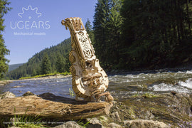 The UGears 3D Hurdy-Gurdy is a beautifully crafted DIY mechanical model made of wood, showcasing intricate gears and an elegant design. It is prominently displayed beside a flowing river, surrounded by lush green trees and mountains. The scenic background enhances the artistic appeal of this musical instrument model, perfect for hobbyists and enthusiasts alike.