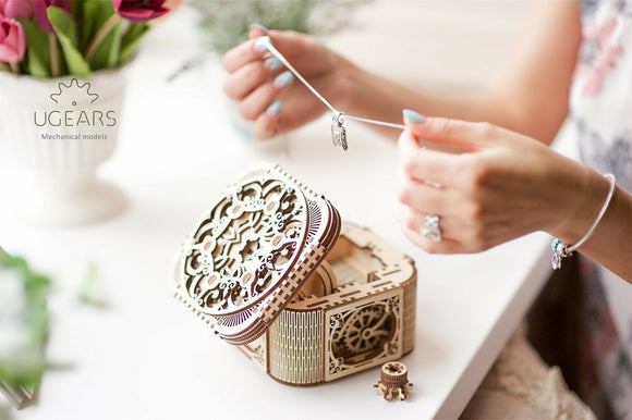 A person assembling the UGears 3D Treasure Box DIY kit, showcasing intricate wooden designs and mechanical elements. The box is partially open, revealing its detailed interior. Surrounding plants and decor add an inviting atmosphere.