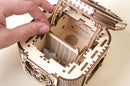 A close-up view of the UGears 3D Treasure Box DIY kit. The intricately designed wooden box features a detailed mechanism with small compartments inside, showcasing its craftsmanship. A hand is placing a coin in the box, highlighting its functional aspect as a treasure container.