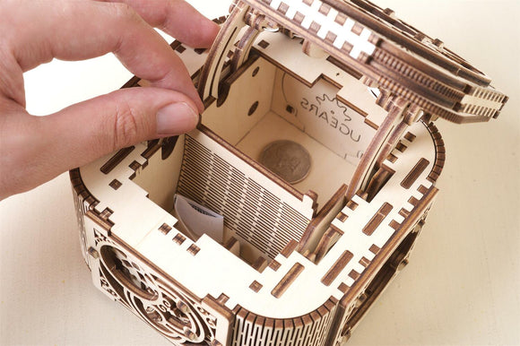 A close-up view of the UGears 3D Treasure Box DIY kit. The intricately designed wooden box features a detailed mechanism with small compartments inside, showcasing its craftsmanship. A hand is placing a coin in the box, highlighting its functional aspect as a treasure container.