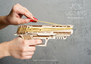 A close-up of a person's hand operating the UGears 3D Wolf-01 Handgun, a mechanical DIY kit made of laser-cut wood. The kit showcases intricate designs and is being loaded with rubber bands for action. The background is a textured gray, emphasizing the craftsmanship of the model.