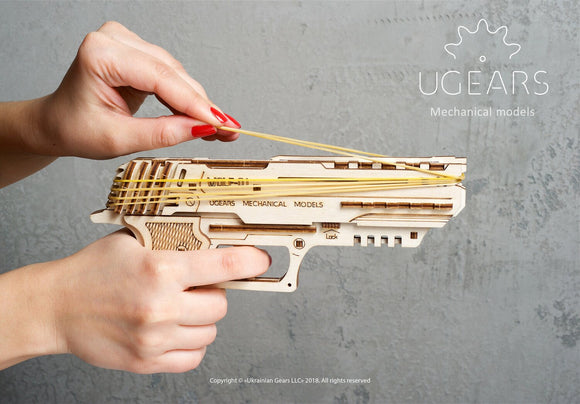 A close-up of a person's hand operating the UGears 3D Wolf-01 Handgun, a mechanical DIY kit made of laser-cut wood. The kit showcases intricate designs and is being loaded with rubber bands for action. The background is a textured gray, emphasizing the craftsmanship of the model.