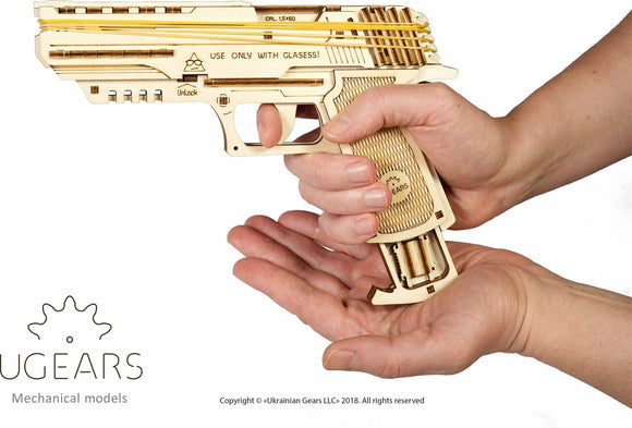 A person holding the UGears 3D Wolf-01 Handgun, a wooden DIY mechanical model kit. The gun features intricate details and allows for hands-on assembly. Warning label visible: 'USE ONLY WITH GLASSES!'