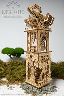 Detailed view of the UGears 3D Archballista-Tower DIY kit, showcasing its intricate wooden design and mechanical components. The model features a tall tower with decorative elements, an archery mechanism at the top, and visible gears, all crafted from high-quality plywood. Set against a natural background with moss and stones, this model combines art and engineering for a delightful building experience.