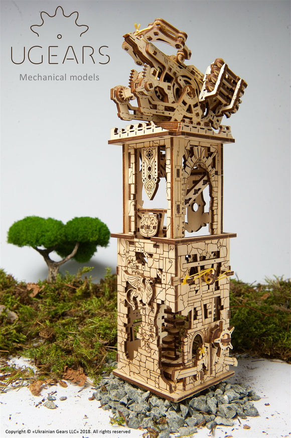 Detailed view of the UGears 3D Archballista-Tower DIY kit, showcasing its intricate wooden design and mechanical components. The model features a tall tower with decorative elements, an archery mechanism at the top, and visible gears, all crafted from high-quality plywood. Set against a natural background with moss and stones, this model combines art and engineering for a delightful building experience.
