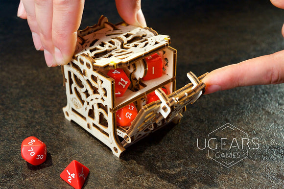 A person is using their finger to open the intricate wooden UGears 3D Dice Keeper. The elegant design showcases its compartments filled with vibrant red dice, while additional dice are placed on a dark surface nearby.