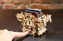 The UGears 3D Deck Box is a beautifully crafted wooden storage solution for cards. Featuring intricate laser-cut designs, it showcases a finger gently pressing on the box, highlighting its mechanical elements and unique construction. The box stands on a dark wooden table, with a backdrop of a rustic brick wall, emphasizing its artistic appeal and functionality as both a decorative piece and practical organizer.