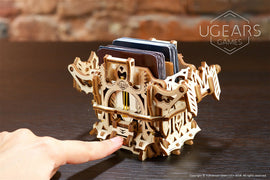 The UGears 3D Deck Box is a beautifully crafted wooden storage solution for cards. Featuring intricate laser-cut designs, it showcases a finger gently pressing on the box, highlighting its mechanical elements and unique construction. The box stands on a dark wooden table, with a backdrop of a rustic brick wall, emphasizing its artistic appeal and functionality as both a decorative piece and practical organizer.