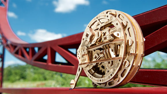 The UGears 3D Monowheel DIY kit displayed outdoors. It features intricate wooden components, showcasing gears and mechanisms, set against a vibrant blue sky and a red roller coaster track. The design emphasizes both aesthetic beauty and mechanical ingenuity, perfect for hobbyists and creators.