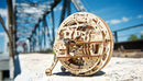 A detailed, wooden 3D puzzle model of a monowheel, intricately designed by UGears 3D. The model showcases moving parts and unique engineering, set against an industrial bridge backdrop under a bright blue sky.