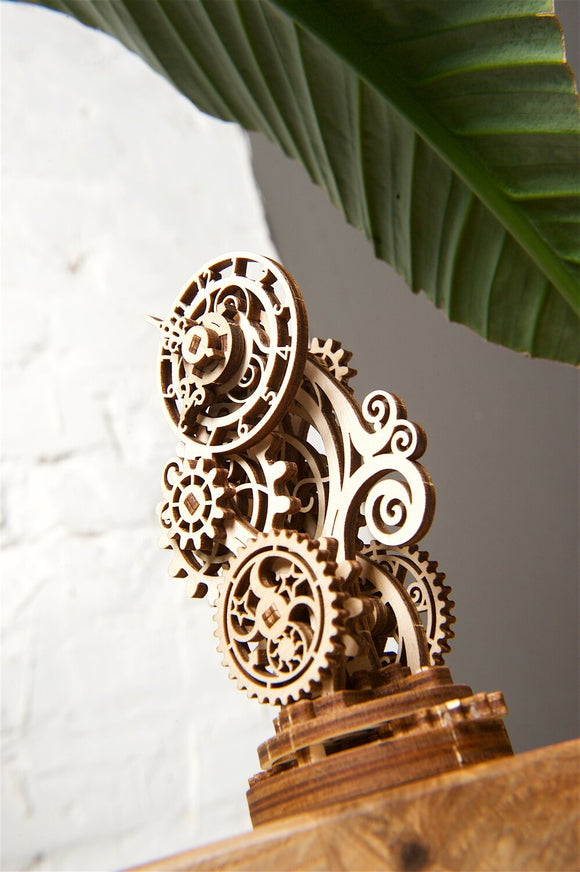 A detailed wooden model of a steampunk clock with intricate gears and decorative elements. The clock is crafted from light wood and showcases various mechanical features, standing on a sturdy base. The background includes a green leaf and a textured wall, adding to the aesthetic.