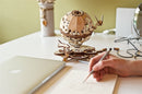 The UGears 3D Globe, a detailed DIY kit made of wood, featuring intricate gears and a globe structure, displayed on a workspace next to a laptop and sketchbook. A hand holds a pencil, emphasizing the creative environment.