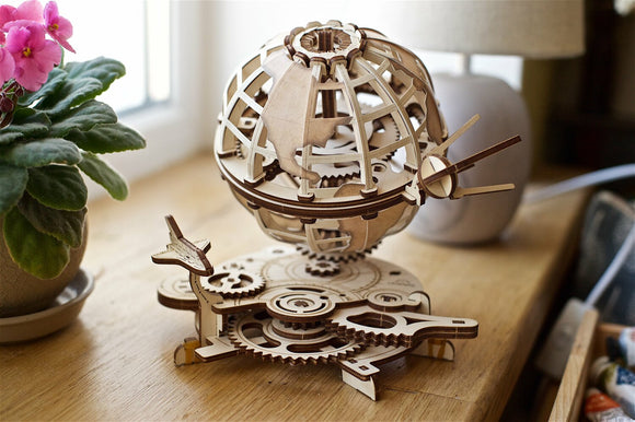 A beautifully crafted UGears 3D globe model showcasing intricate wooden gears and a globe structure. The globe is resting on a circular base surrounded by a potted plant and a lamp, highlighting its stunning design and engineering.